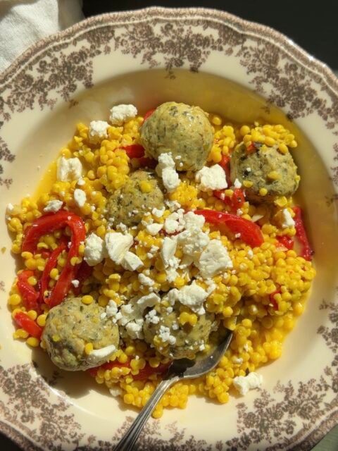 A bowl of pearl couscous mixed with red bell peppers, topped with Greek meatballs and crumbled feta cheese, with a silver spoon resting in the bowl.