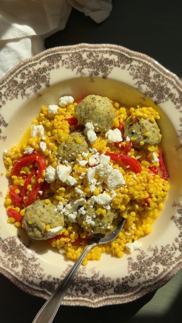 A bowl of pearl couscous mixed with red bell peppers, topped with Greek meatballs and crumbled feta cheese, with a silver spoon resting in the bowl.