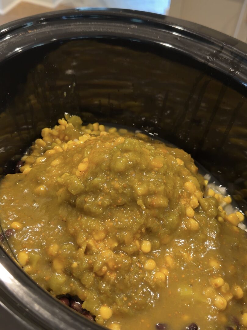 A slow cooker filled with black beans, corn, and a thick layer of green salsa.