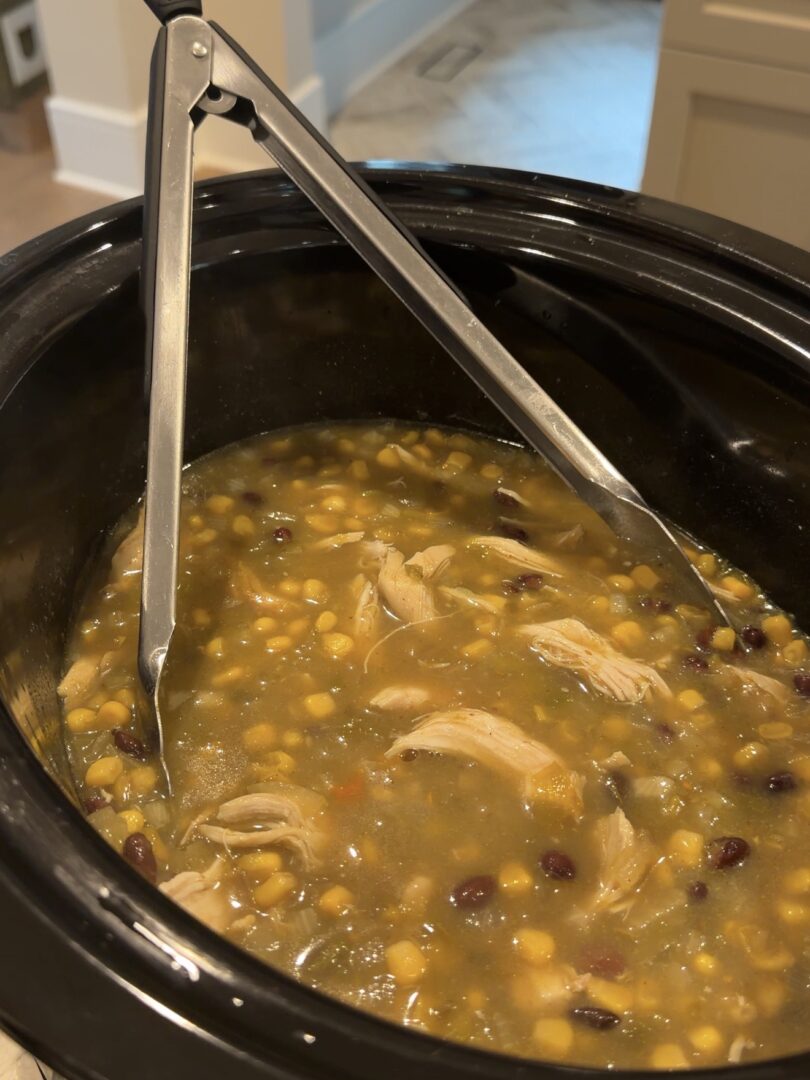 A slow cooker filled with chicken, corn, and black bean soup, with metal tongs resting inside.