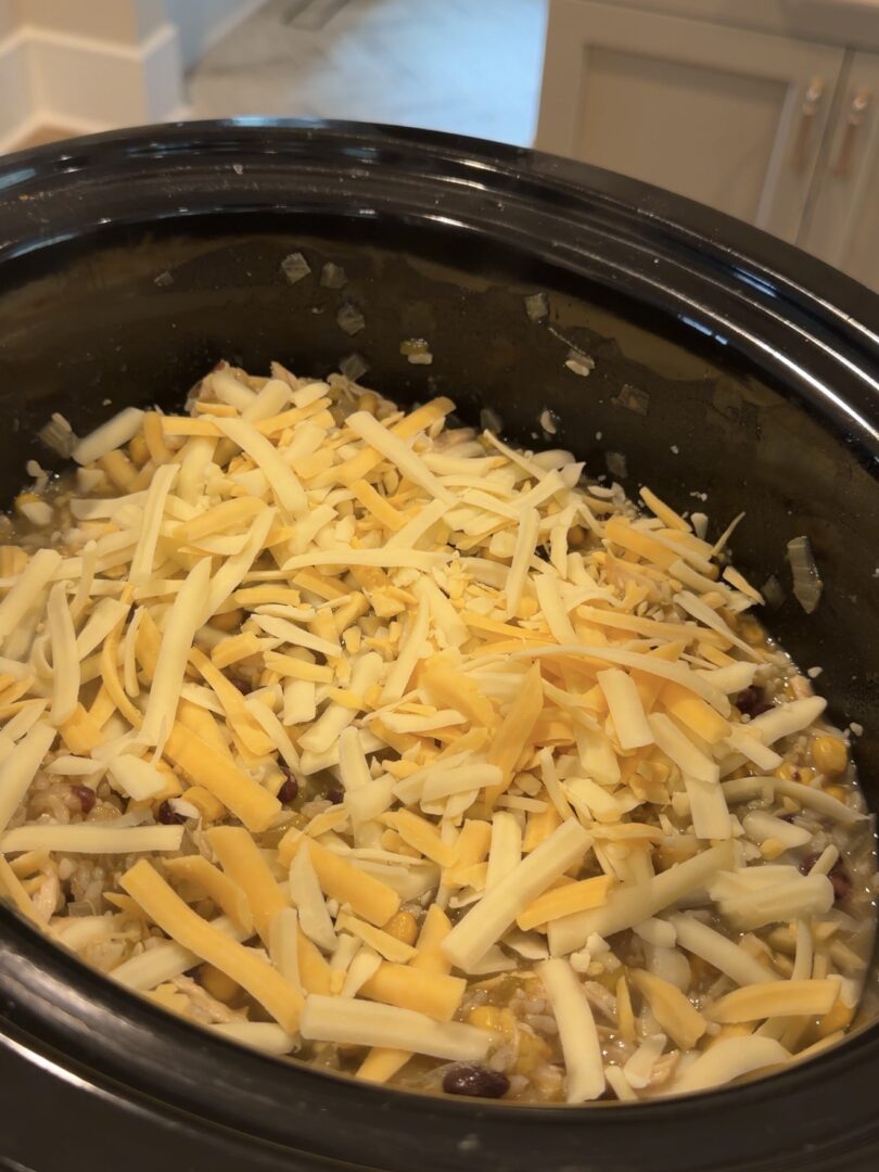 A slow cooker filled with a mixture of beans, corn, and grains topped with a thick layer of shredded cheddar cheese.