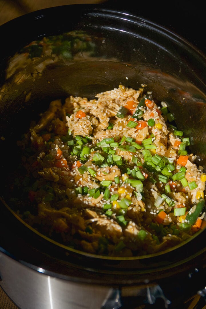 A slow cooker filled with chicken and vegetable fried rice topped with sesame seeds and chopped green onions.
