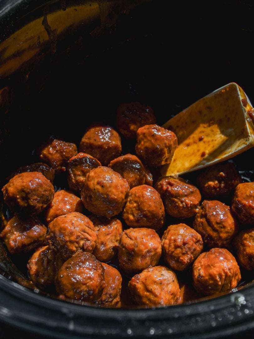 Meatballs in a slow cooker coated in a glossy, dark red sauce with a spatula resting in the pot.
