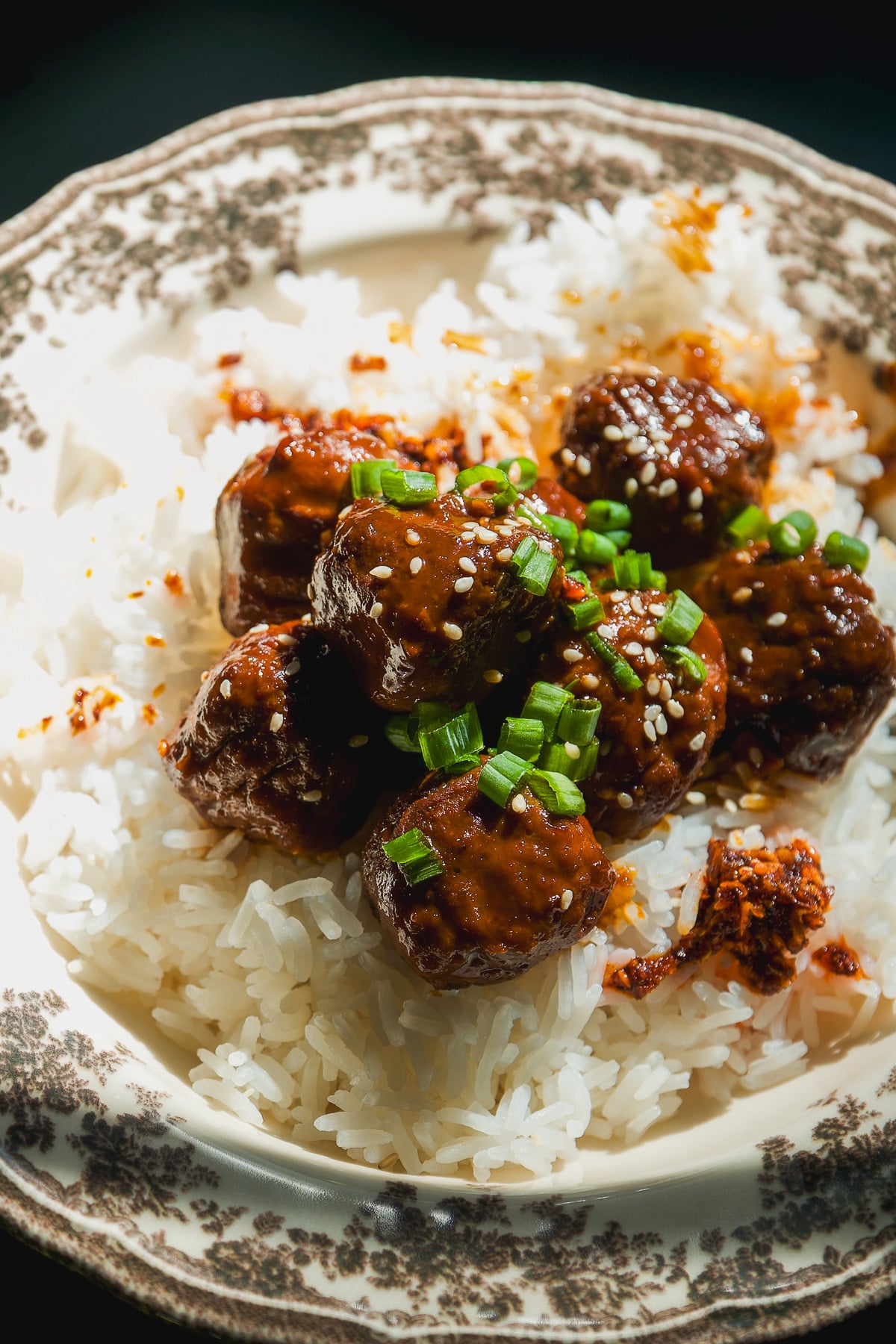 Meatballs glazed in dark sauce, topped with sesame seeds and chopped green onions, served over a bed of white rice in a patterned bowl.