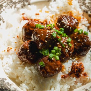 Meatballs in a dark glaze topped with sesame seeds and green onions, served over a bed of white rice in a patterned bowl.