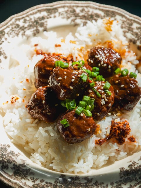 Meatballs glazed in sauce, topped with sesame seeds and chopped green onions, served over a bowl of white rice.