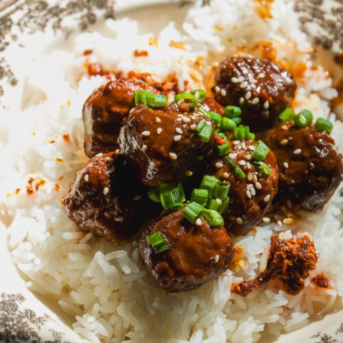 Meatballs glazed in sauce, topped with sesame seeds and chopped green onions, served over a bowl of white rice.