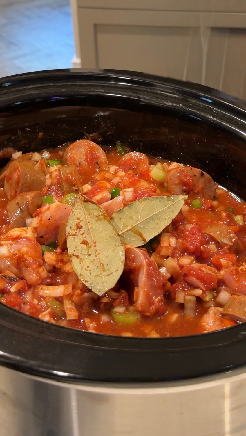 A slow cooker filled with a stew of sliced sausage, chopped vegetables, and tomato sauce, topped with two bay leaves.