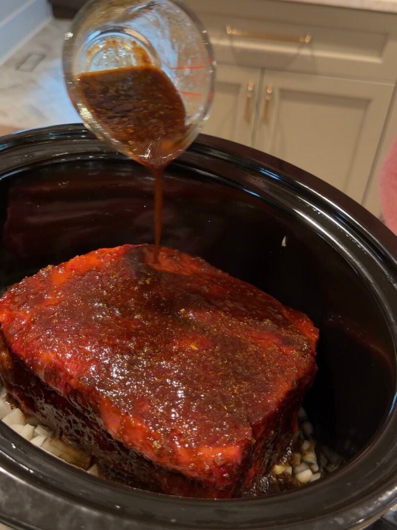 A hand pours a dark brown sauce from a measuring cup over a seasoned beef roast inside a slow cooker.
