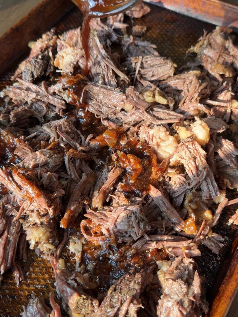 Sauce being poured from a glass container over a pan of shredded beef.