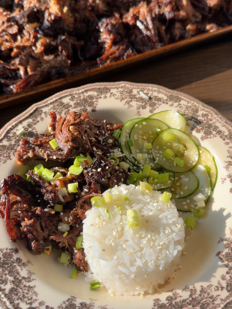 A plate with shredded beef, a mound of white rice, and sliced cucumbers, garnished with sesame seeds and green onions, with a baking sheet of shredded beef in the background.