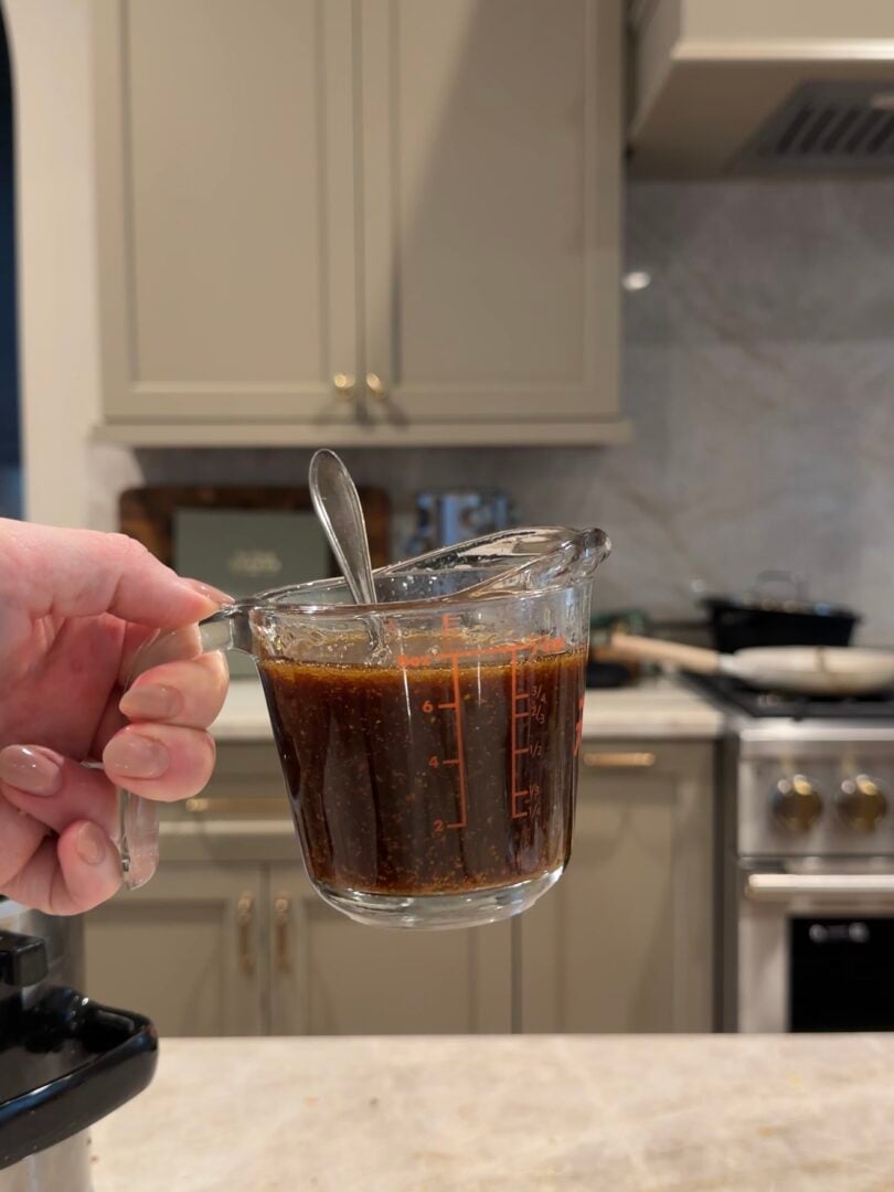 A hand holds a glass measuring cup filled with a dark, textured sauce and a metal spoon.