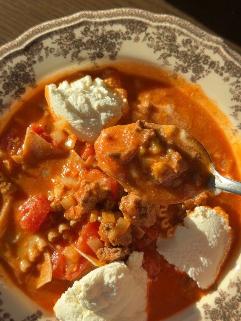 A spoonful of lasagna soup with chunks of meat and tomato, served in a patterned bowl topped with dollops of ricotta cheese.