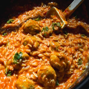 Creamy tomato orzo with meatballs and greens in a slow cooker, with a wooden spoon resting in the dish.