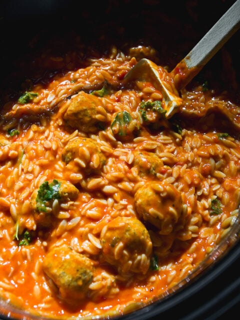 Crockpot meatballs and orzo in a creamy tomato sauce with chopped greens, served in a slow cooker with a wooden spoon resting in the center.