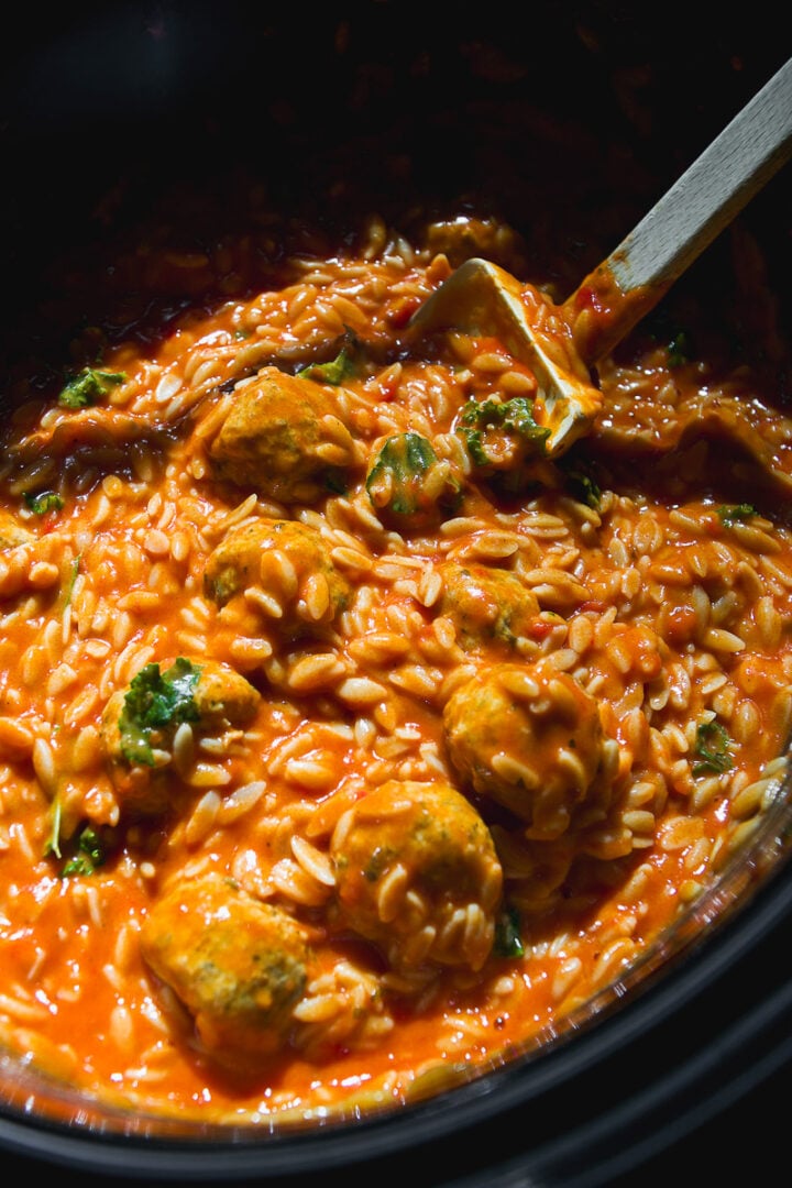 Crockpot meatballs and orzo in a creamy tomato sauce with chopped greens, served in a slow cooker with a wooden spoon resting in the center.
