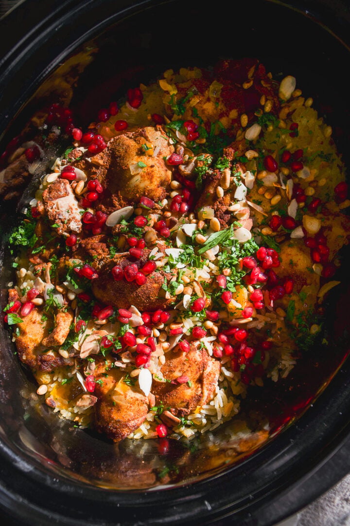 A slow cooker filled with chicken and rice topped with pomegranate seeds, nuts, and fresh herbs.