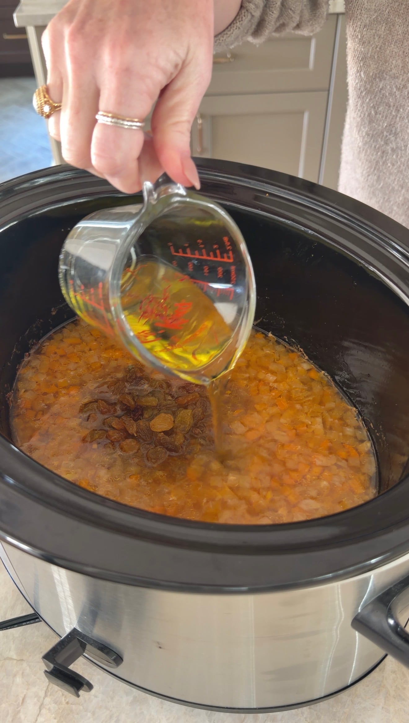 A hand pours liquid from a measuring cup into a slow cooker containing a mixture of chopped vegetables and raisins.