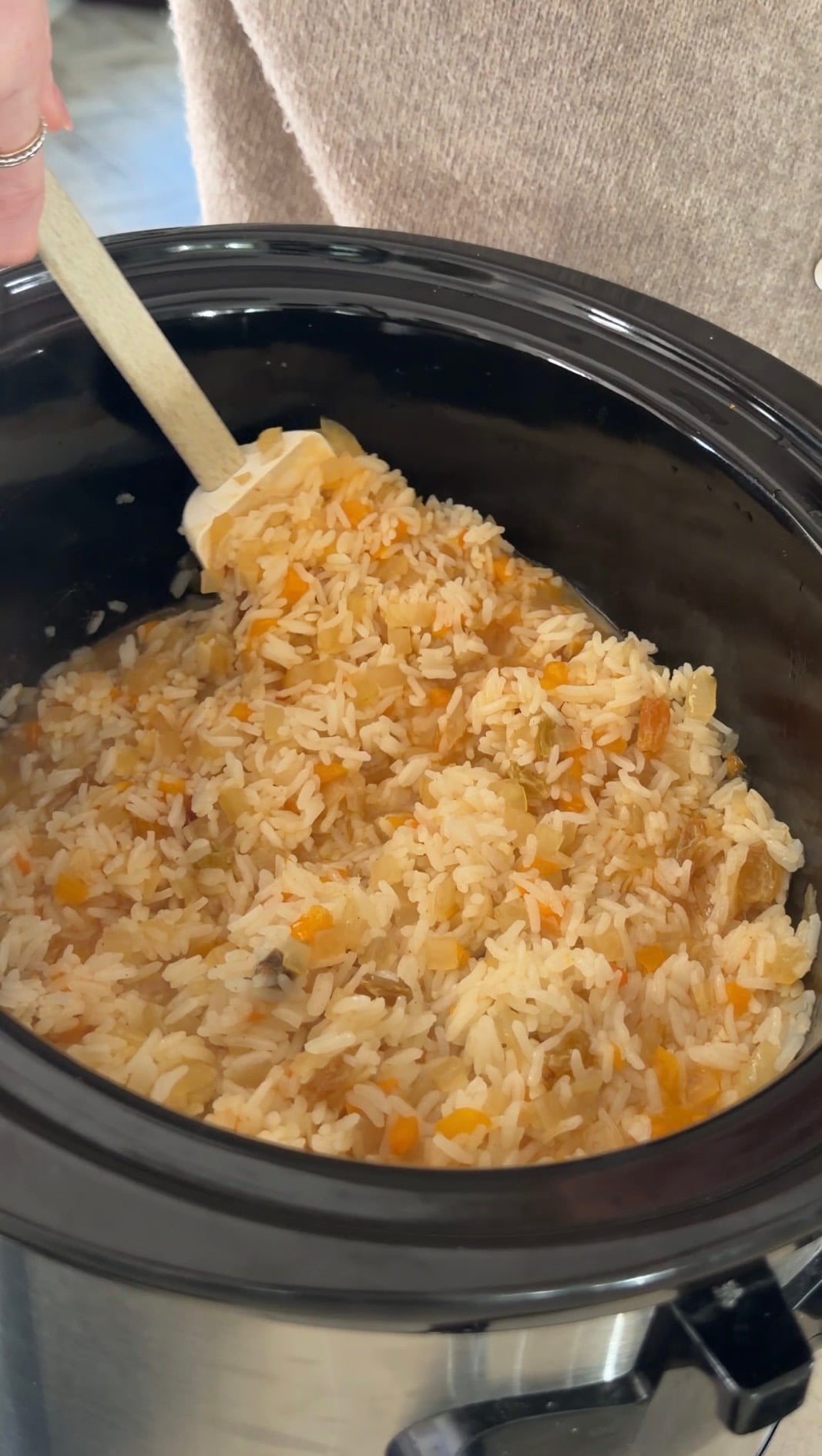 A hand uses a white silicone spatula to stir cooked rice mixed with diced vegetables in a slow cooker.