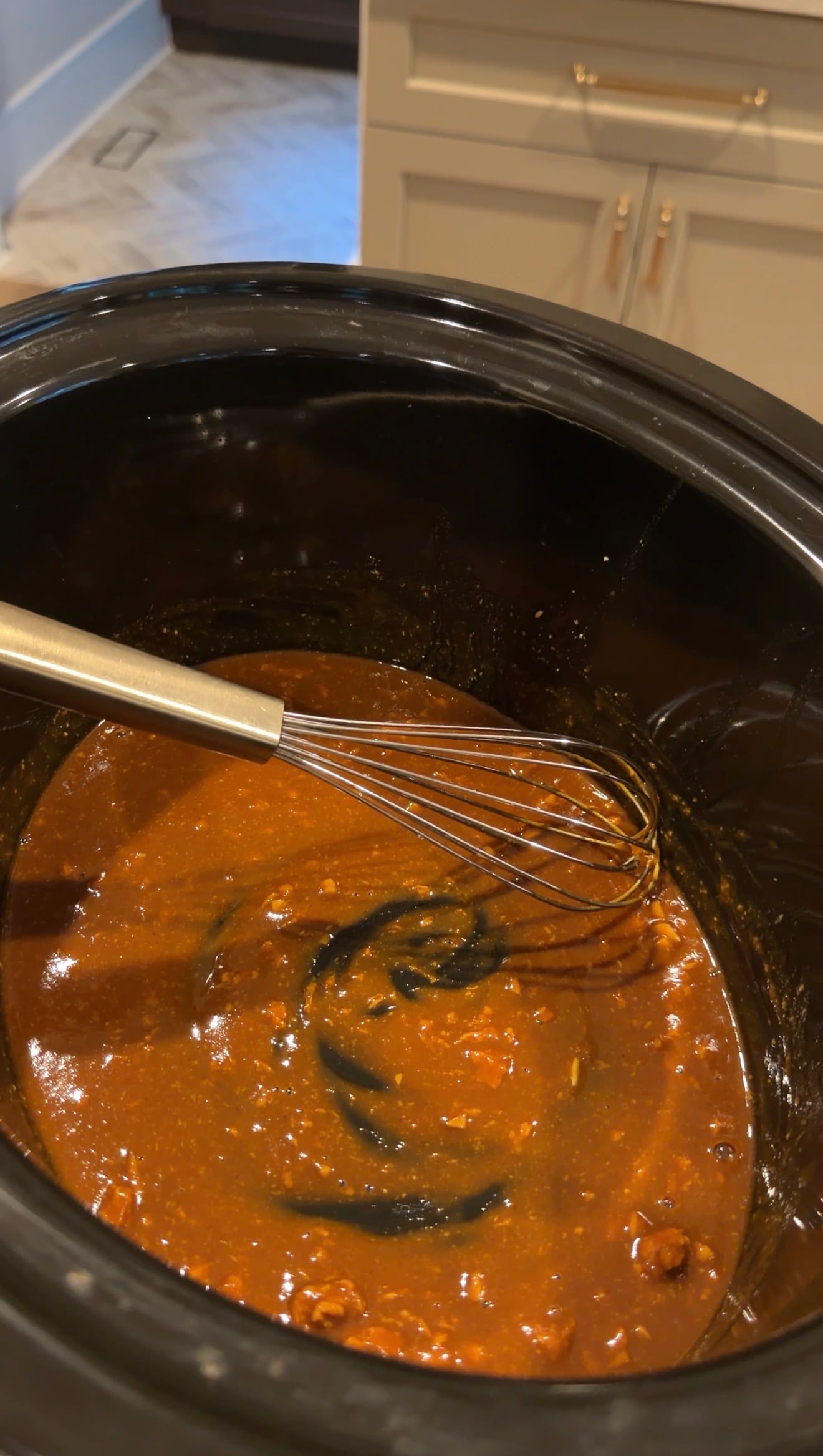 A metal whisk stirs a thick, reddish-brown sauce in a black slow cooker.