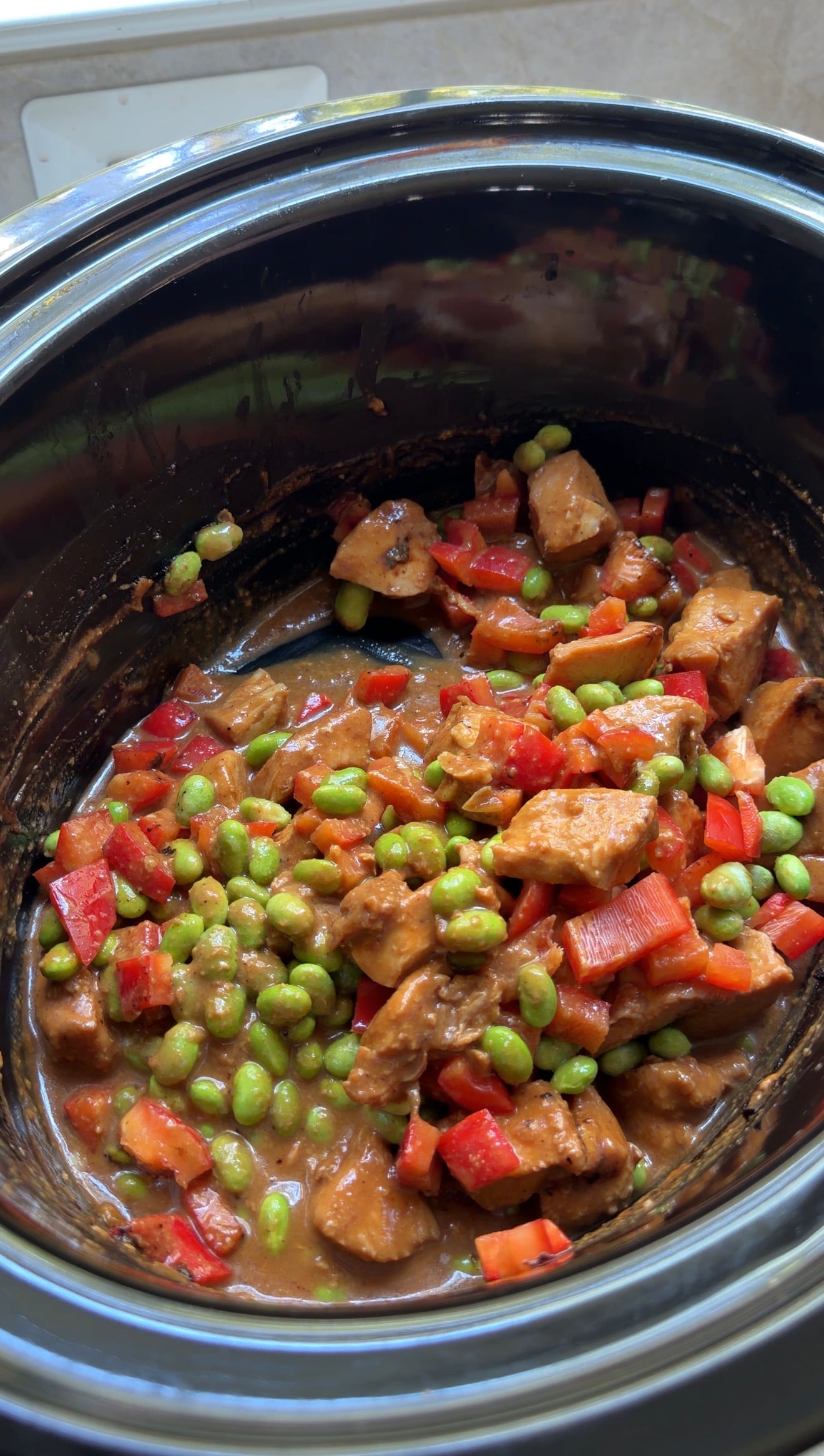 A slow cooker filled with a stew of chicken, edamame, and red bell peppers in a brown sauce.