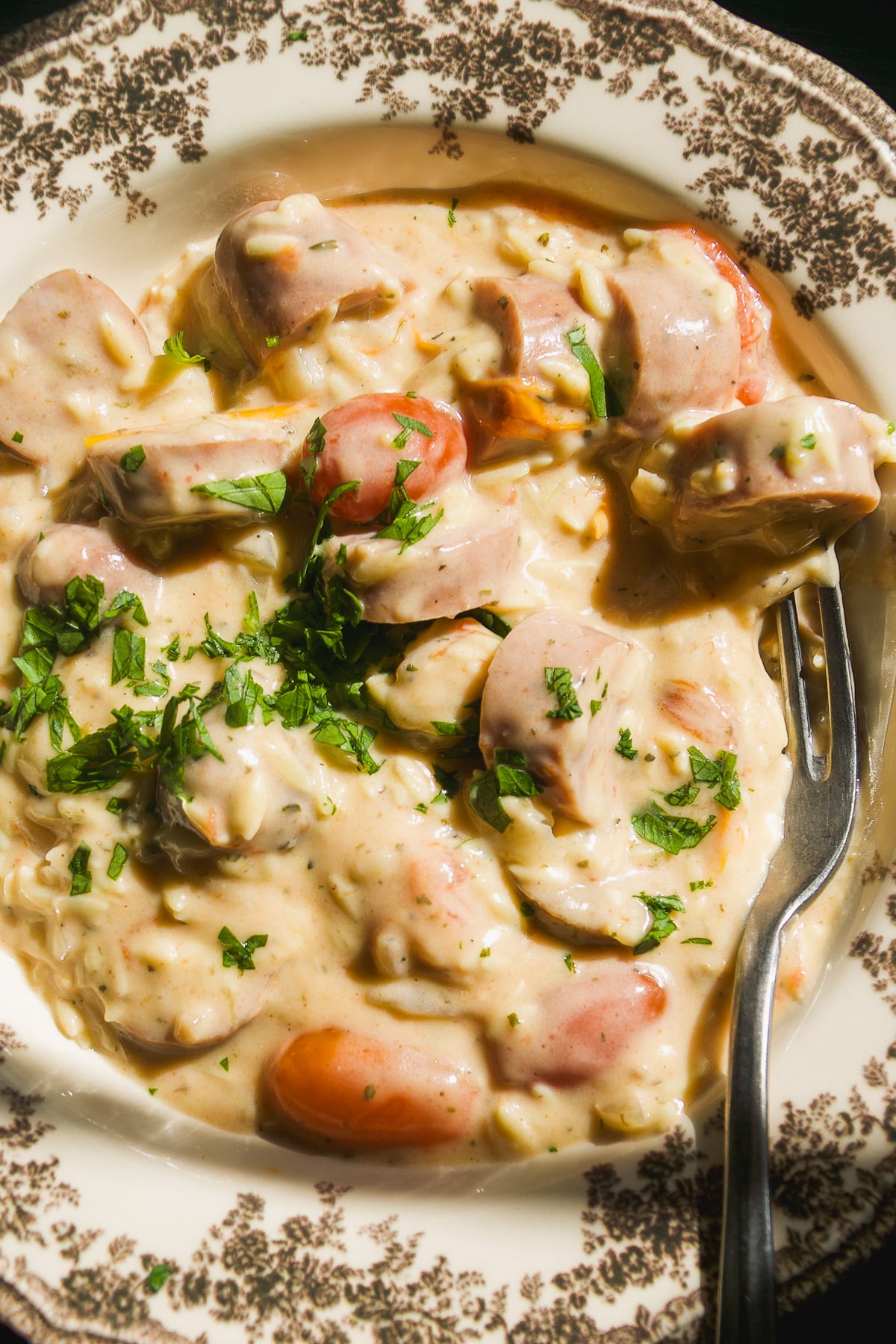 A bowl of creamy pasta with sliced sausages, garnished with fresh herbs and served with a fork.