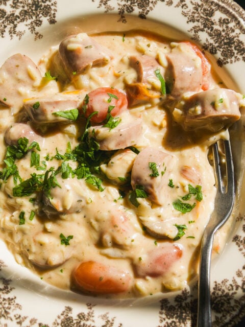 Creamy pasta with sliced sausage and herbs in a patterned bowl, with a fork resting on the side.