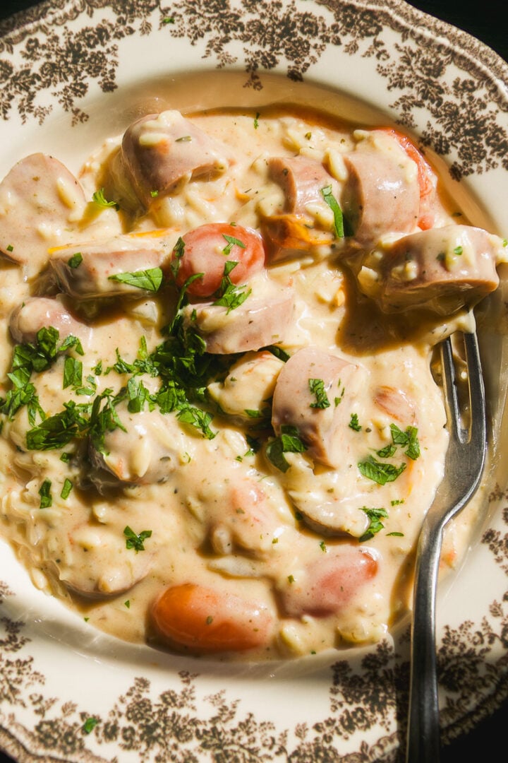 Creamy pasta with sliced sausage and herbs in a patterned bowl, with a fork resting on the side.