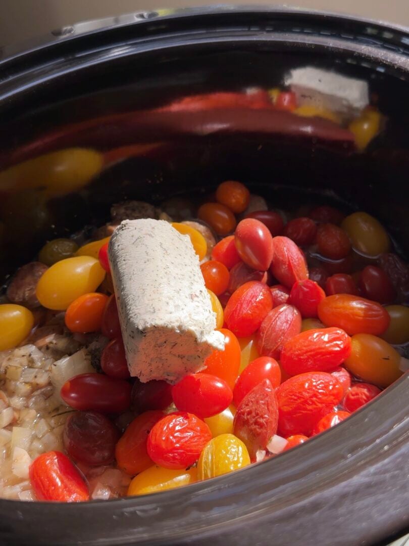 A block of herb-speckled cream cheese sitting atop a mixture of fresh cherry tomatoes and chopped onions in a slow cooker.