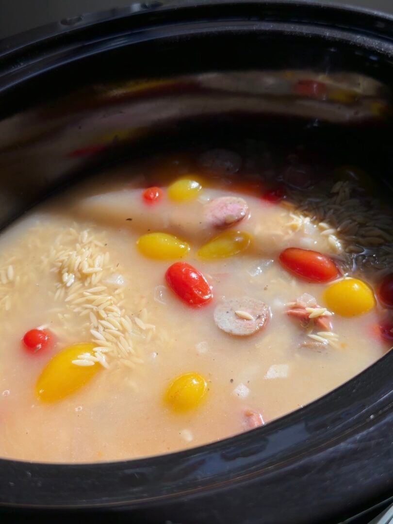 Slow cooker filled with a creamy broth containing orzo pasta, cherry tomatoes, and sliced sausage.