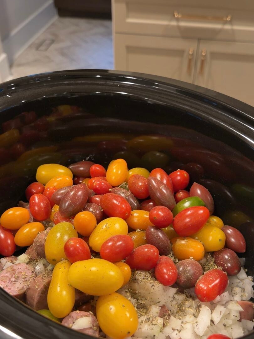 A slow cooker filled with colorful cherry tomatoes, chopped onions, and sausage seasoned with herbs.