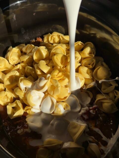 Cream being poured over raw tortellini in a slow cooker.