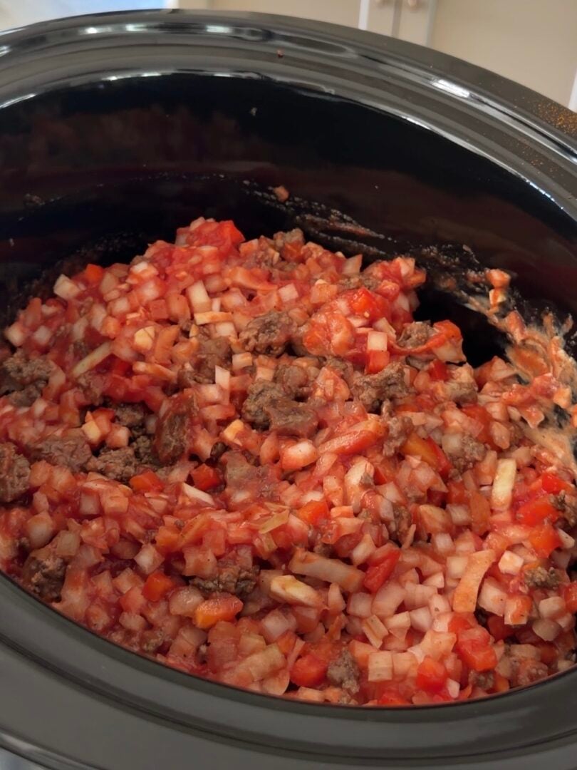 A slow cooker filled with a mixture of ground meat, diced white onions, and red bell peppers in a tomato-based sauce.