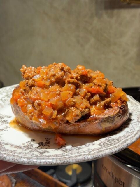 A baked sweet potato topped with a savory ground meat and tomato sauce on a patterned plate.