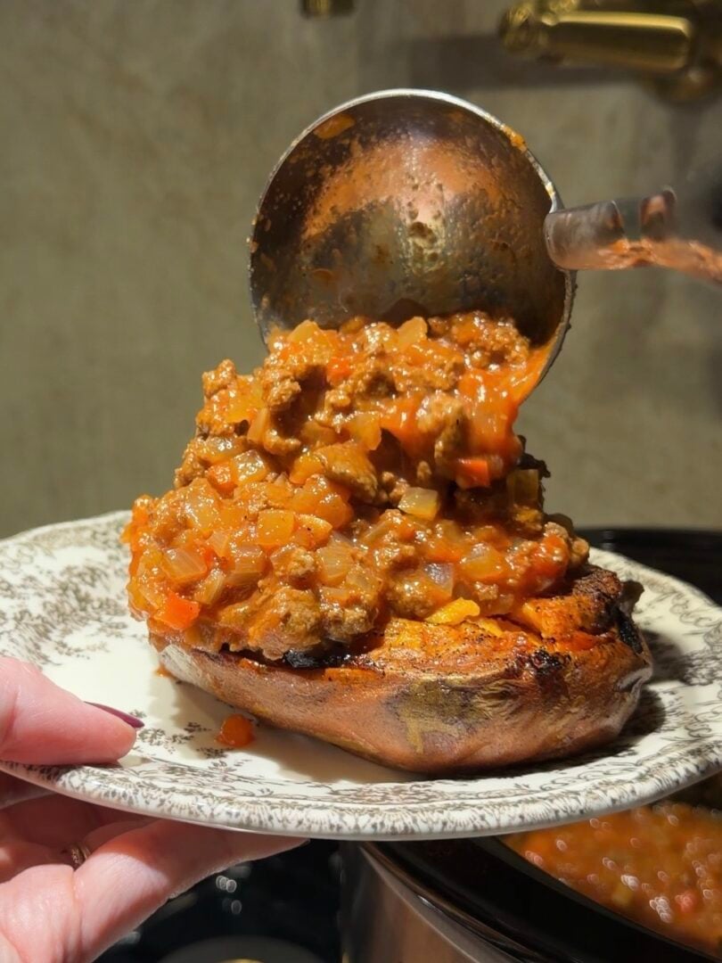 A hand holds a plate as a ladle pours savory meat and vegetable sauce over a baked sweet potato.