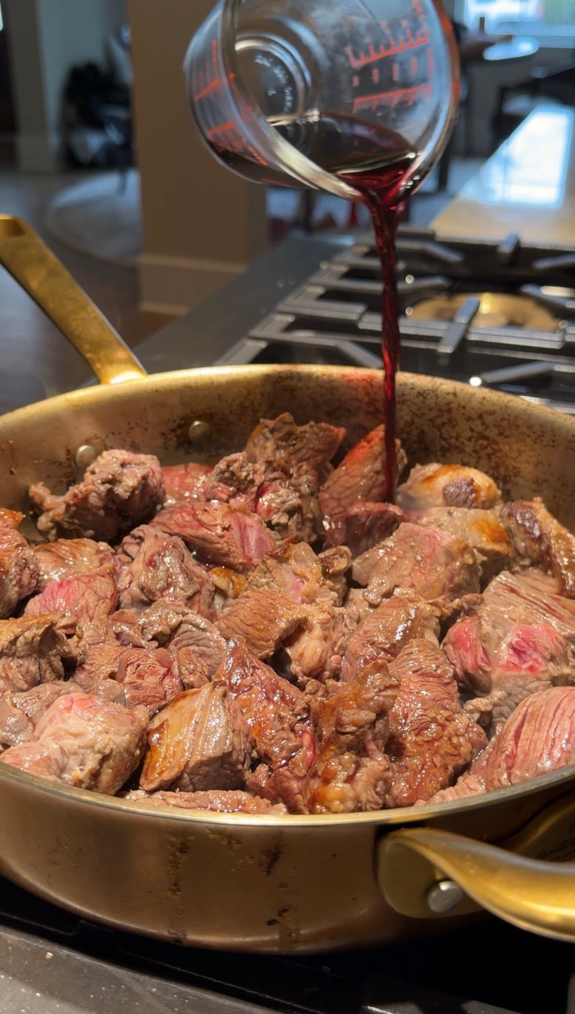 Red wine being poured from a measuring cup into a pan of searing beef cubes for a crockpot steak and potatoes recipe.