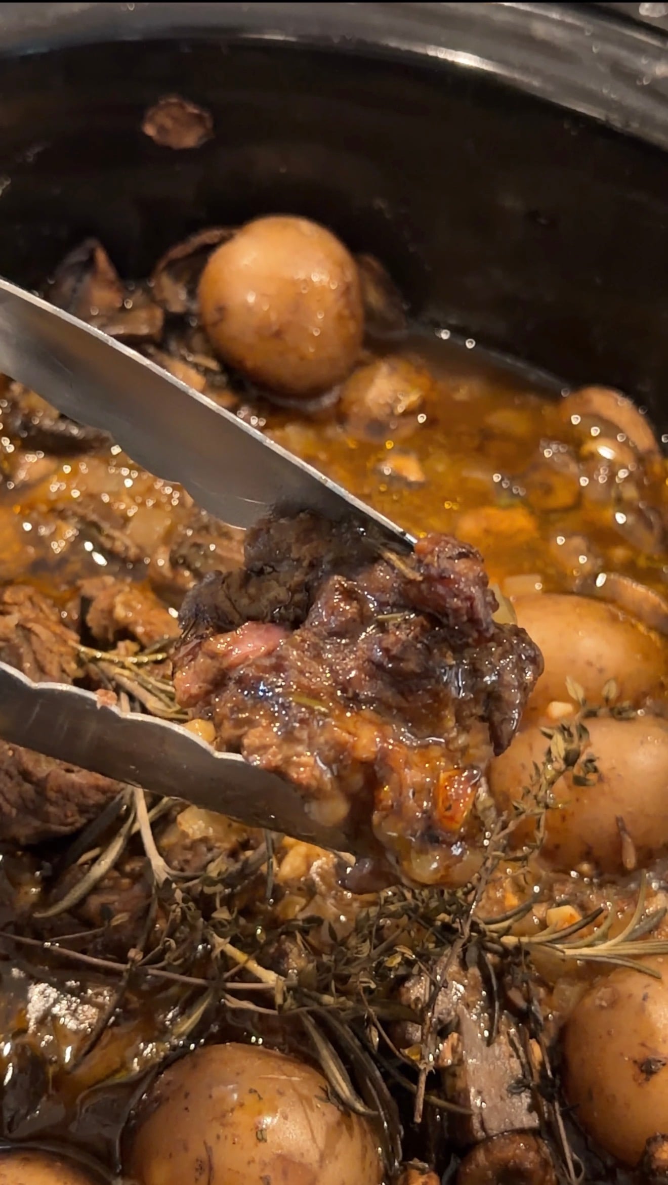 Metal tongs lifting a piece of Crockpot steak and potatoes from a slow cooker filled with herbs and savory liquid.