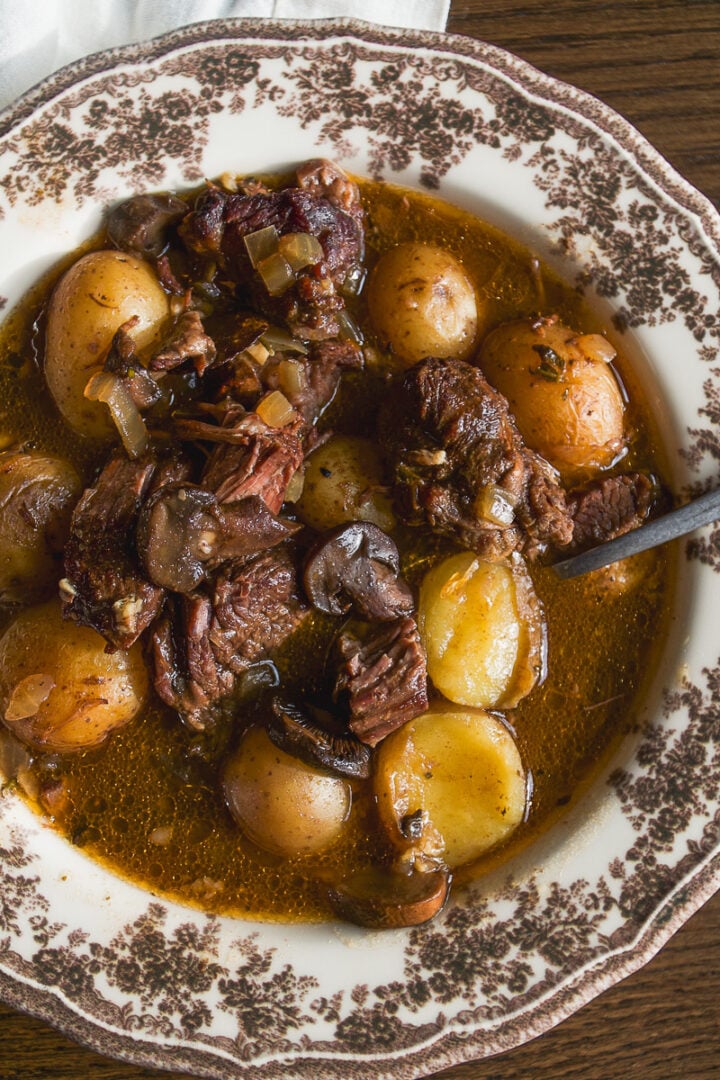 A bowl of beef stew with chunks of meat, mushrooms, and whole baby potatoes in a dark broth.