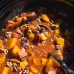 A slow cooker filled with a stew of cubed squash, black beans, and ground meat in a tomato-based broth, with a serving spoon resting inside.