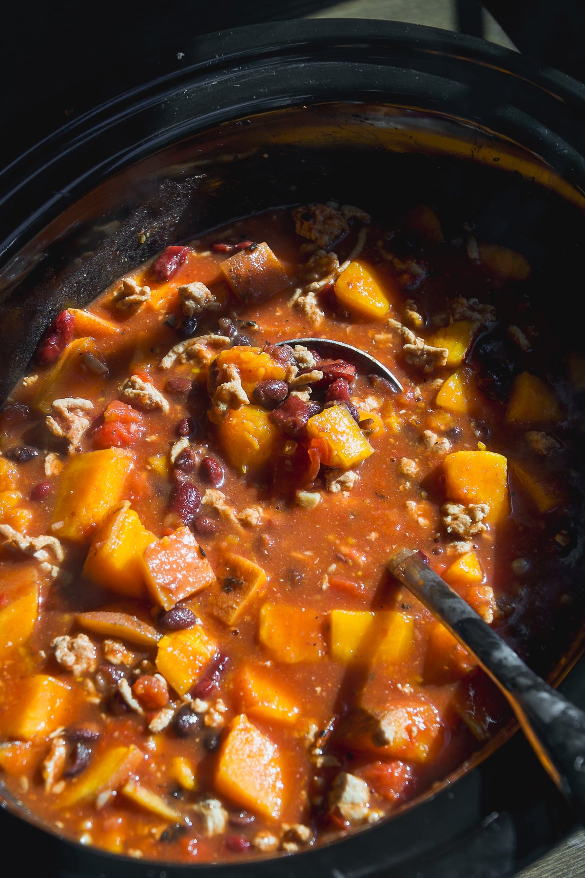A slow cooker filled with a stew of cubed squash, black beans, and ground meat in a tomato-based broth, with a serving spoon resting inside.