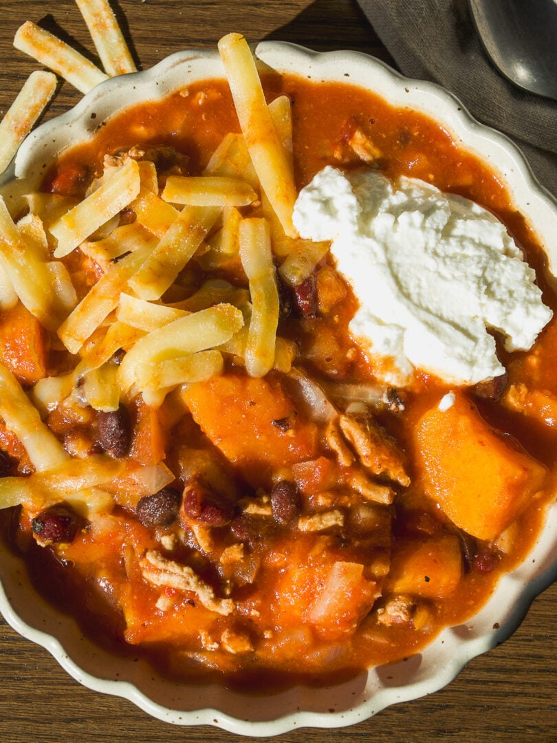 A bowl of chili with sweet potato, beans, and meat, topped with shredded cheese and a dollop of sour cream.