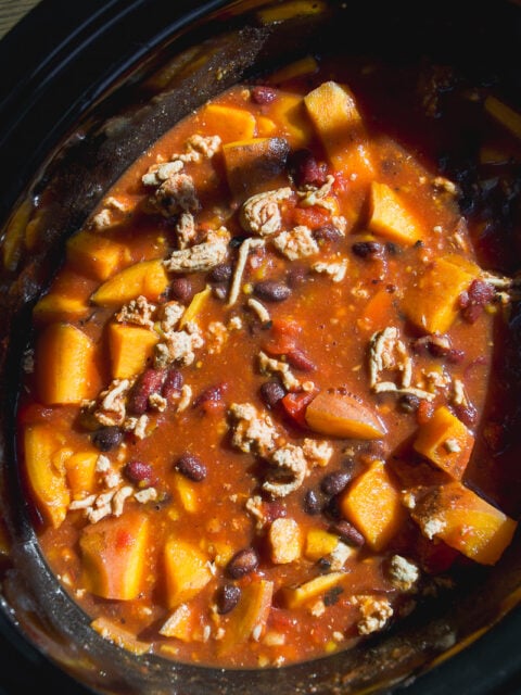 A slow cooker filled with a stew of cubed squash, black beans, and ground meat in a tomato-based sauce.