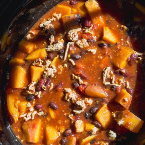 A slow cooker filled with a stew of cubed squash, black beans, and ground meat in a tomato-based sauce.