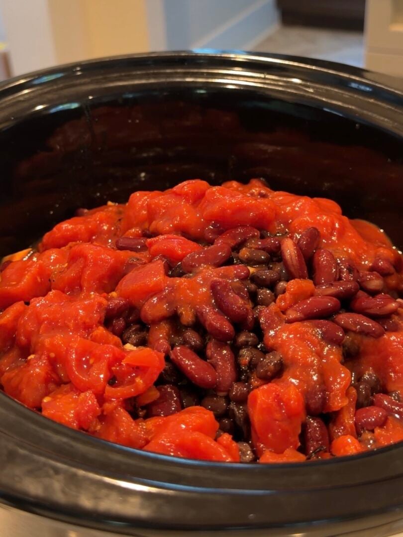 A slow cooker filled with a mixture of red kidney beans, black beans, and diced tomatoes.