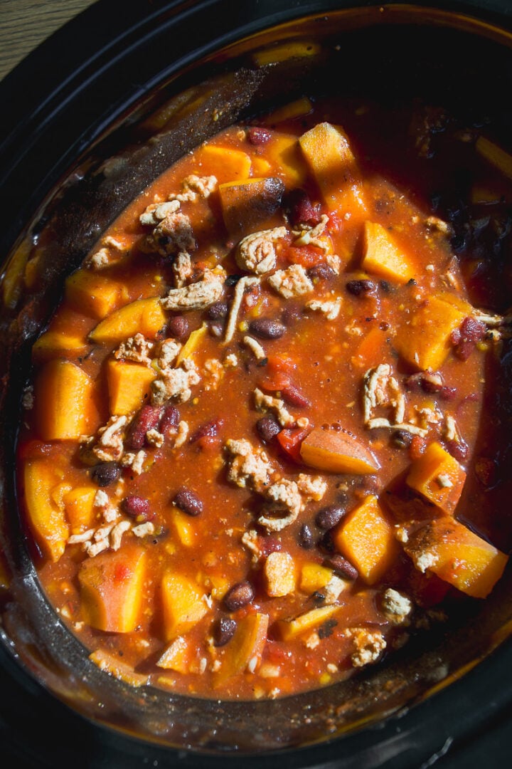 A slow cooker filled with a stew of cubed squash, black beans, and ground meat in a tomato-based sauce.