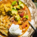 A bowl of chili topped with avocado chunks, shredded cheese, tortilla chips, and a dollop of sour cream.