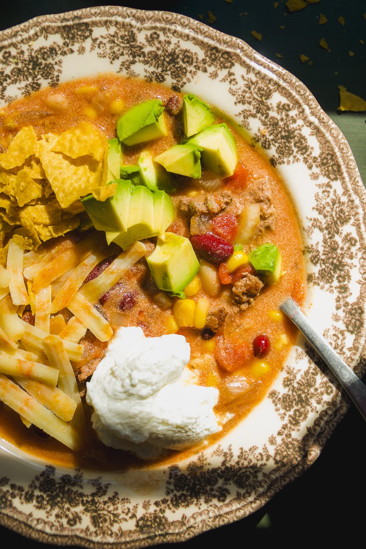 A bowl of chili topped with avocado chunks, shredded cheese, tortilla chips, and a dollop of sour cream.