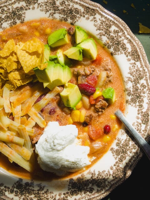 A bowl of chili topped with diced avocado, shredded cheese, a dollop of sour cream, and tortilla chips.