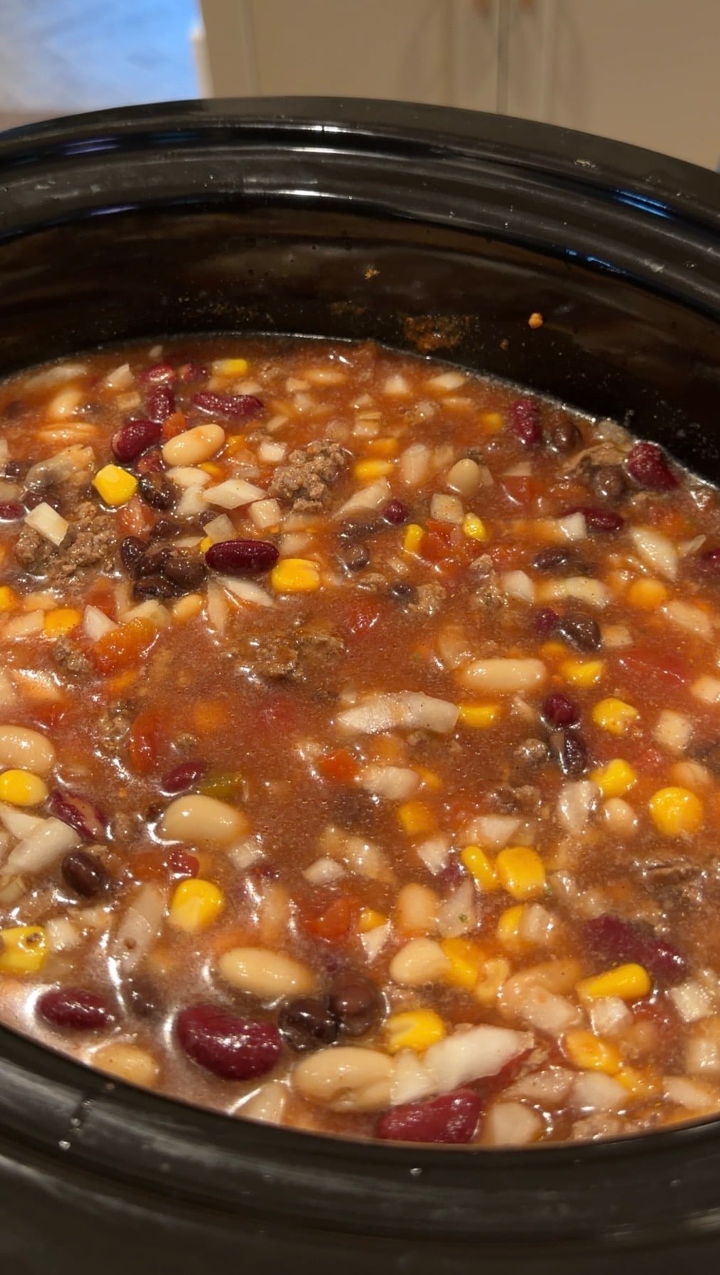 A slow cooker filled with a chunky soup containing beans, corn, and ground meat in a tomato-based broth.