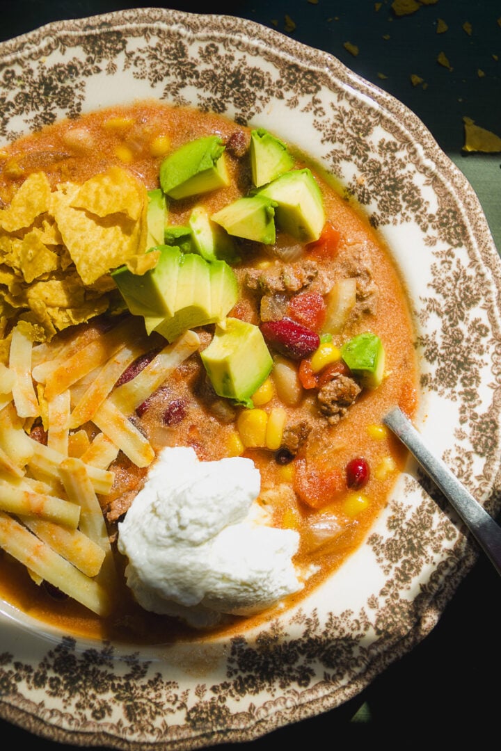 A bowl of chili topped with diced avocado, shredded cheese, a dollop of sour cream, and tortilla chips.